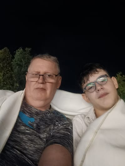 Two individuals, an adult male and a juvenile male, are seated close together, possibly on a couch or lounge chairs, wrapped in white blankets. They are both wearing glasses and appear to be outdoors at night, with dark foliage visible in the background. The adult male is on the left, wearing a striped t-shirt with a blue graphic. The juvenile male is on the right, wearing a white collared shirt. The image does not contain enough information to determine a specific location in Tserovani, Georgia.