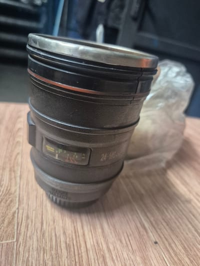 The image shows a close-up of a camera lens, which appears to be a telephoto or zoom lens. The lens is black and has several rings and markings. There is a distance scale visible, with markings for feet (ft) and meters (m), including a "MACRO" setting. The lens is resting on a light-colored wooden surface, likely a floor or table. In the background, a blurry, light-colored object, possibly a bag or cloth, is visible. The lighting suggests an indoor setting, possibly during the day. No people are visible in the image. Visible text on the lens includes "24-105" and "IMAGE STABILIZER". The context provided indicates the location is Balqaş, Kazakhstan.