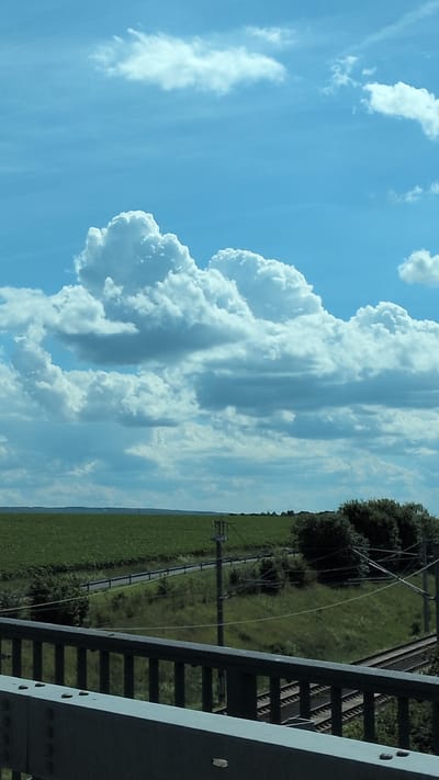 This is a vertical, landscape photograph taken on a sunny day with a bright blue sky and scattered white, puffy clouds. The image is split between the sky and a field with power lines. The foreground is dominated by a metal structure, possibly a bridge or railway overpass, running diagonally from the bottom right to the top left of the frame. The structure is a dark gray color and features a railing with vertical bars.

To the left of the structure, a vast green field stretches towards the horizon. Several utility poles with wires crisscross the field, suggesting the presence of electrical infrastructure. Trees are visible in the distance along the edge of the field. The overall scene evokes a sense of rural openness. The lighting and cloud formations indicate it is likely daytime, possibly late morning or afternoon. There is no visible text in the image.