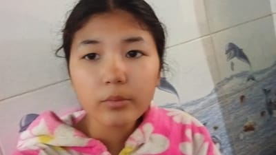 A young girl with dark hair is shown in a close-up selfie. She is wearing a pink and white patterned robe. The background appears to be a tiled wall with a mural depicting dolphins and waves. The girl's face is illuminated from the front, and her gaze is directed towards the camera. The scene suggests an indoor setting, possibly a bathroom or dressing area. The image is taken in Uchkun, Kyrgyzstan.