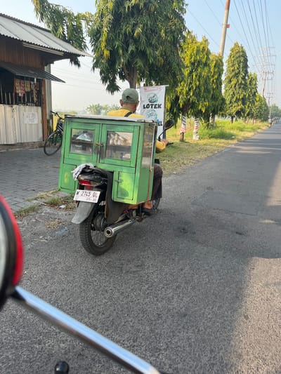 A man wearing a yellow shirt and a green cap rides a motorcycle with a green wooden food cart attached to the back. The cart has glass doors displaying food items and a license plate that reads "W 2745 QL". The motorcycle is on a paved road in what appears to be a semi-rural area, with trees lining the road and a small building with goods for sale on the left. In the background, power lines and transmission towers are visible. The scene is set in Karangtanjung, Indonesia, during daylight hours.