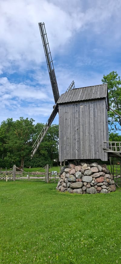 An old, weathered wooden windmill stands in a grassy field under a partly cloudy blue sky. The windmill has a square tower, a pitched roof, and one large, unfinished wooden blade angled towards the sky. The base of the windmill is a circular mound of large, rounded stones. A wooden walkway leads to an entrance on the right side of the tower. A wooden fence with multiple horizontal rails encloses a section of the field to the left of the windmill. Lush green trees form a backdrop behind the fence and windmill. The field is covered in green grass dotted with small white wildflowers. The scene appears to be set in Angla, Estonia, possibly at the Angla Heritage Mill complex, known for its windmills. There are no people or animals visible in the image.