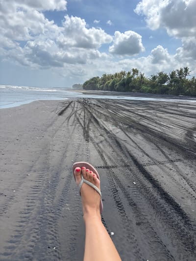 The image shows a beach scene in Kerambitan, Indonesia. The beach has black sand marked with tire tracks. The ocean meets the sand on the left of the frame. Vegetation lines the beach in the background. The sky is visible above with a combination of blue sky and white clouds. A fair-skinned person's foot wearing a silver sandal with red painted toenails is visible on the right side of the frame.