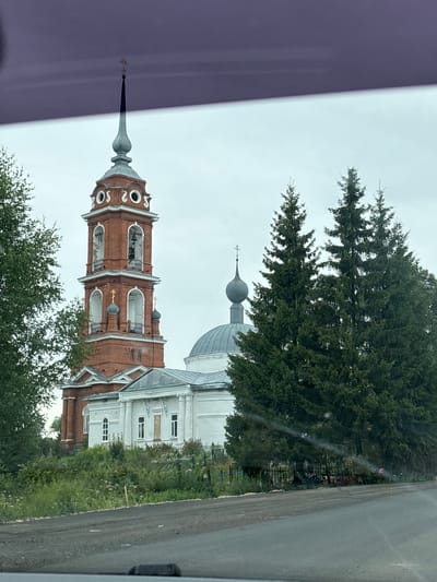 In Novyy Nekouz, Russia, a tall, red brick bell tower with white arched openings and a gray spire topped with a cross stands prominently. Adjacent to the bell tower is a white church with a domed roof and columns. The structures are surrounded by lush green trees, including several tall pine trees, and vegetation. The foreground shows a gray asphalt road. The sky is overcast and light gray.