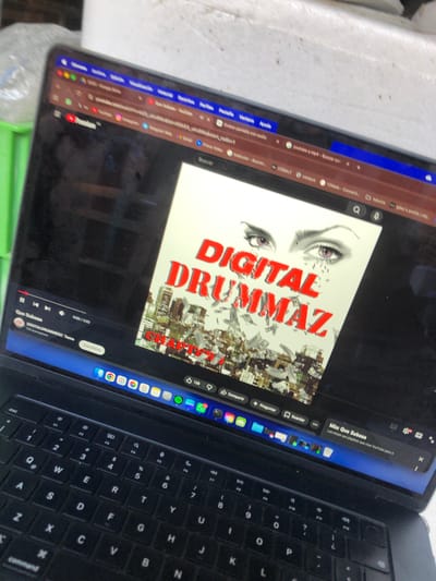 The image shows a laptop screen displaying a YouTube video with the title "DIGITAL DRUMMAZ" in large red letters. The video features an album cover depicting eyes above a cityscape with scattered money. The laptop's keyboard is visible to the right. The laptop is placed on a surface that appears to be part of a table or desk. The environment suggests a casual setting, possibly indoors. There is no information to suggest this image was taken in Beccar, Argentina.