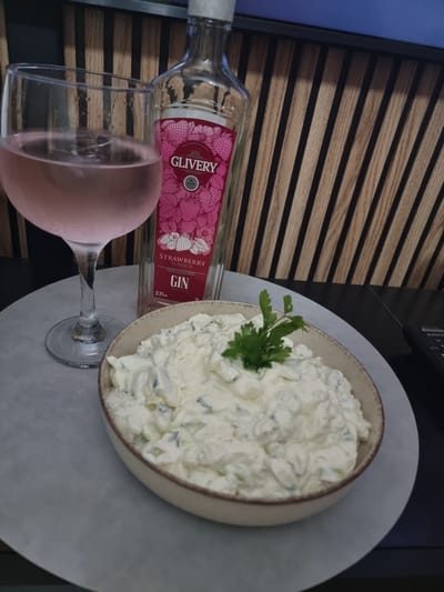 The image captures an indoor still life featuring a bottle of gin, a drink, and a bowl of dip, arranged on a light gray circular placemat atop a surface. The background consists of a wooden slatted wall panel, with a dark electronic device partially visible on the right and a blue surface on the far left.

A clear glass bottle of "GLIVERY STRAWBERRY FLAVOUR GIN" is prominent, with its label displaying strawberries and the text "37.5% vol" and "70cl". Next to the bottle, a martini-style glass holds a light pink beverage, likely prepared with the strawberry gin. To the right, a ceramic bowl is filled with a creamy white dip, which appears to contain finely chopped green ingredients, possibly cucumber or herbs. A fresh sprig of parsley garnishes the center of the dip.

The arrangement suggests a casual setting for enjoying drinks and snacks, indicative of relaxation or social gathering, possibly in a home in Xàtiva, Spain. No people are visible, and the time of day is not discernible.