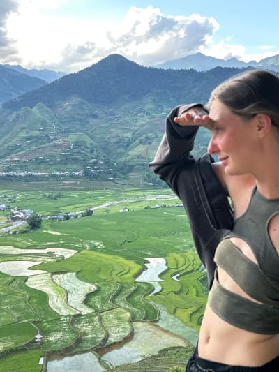A young person, visible from the midriff up and positioned on the right side of the frame, stands on a high vantage point overlooking the Tú Lệ valley in Vietnam. The individual is facing right, with their right hand raised to their forehead, shielding their eyes as they gaze into the expansive landscape. They are wearing a dark grey open zip-up hoodie partially draped over their left shoulder, revealing a dark green, cut-out style crop top. Their midriff and navel are visible. The background presents a vast panoramic view dominated by undulating mountains, some densely covered in green foliage, under a partly cloudy sky. The valley floor is characterized by an intricate pattern of vibrant green terraced rice paddies, many filled with reflective water. A river or stream winds through the valley, bordered by scattered clusters of houses and a visible network of roads ascending the mountain slopes.