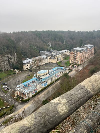 An elevated perspective captures a section of Bagnoles de l'Orne Normandie, France, situated within a densely forested valley under an overcast sky. In the immediate foreground, a weathered grey wooden railing, exhibiting distinct wood grain and lichen growths, extends diagonally from the lower right, partially obscuring the lower portion of the scene. A wire mesh fence is visible directly below the railing.
The central part of the valley contains a complex of buildings. In the mid-ground, a long, low-rise building with light-colored walls and a distinctive light-blue roof, patterned with concentric wave-like designs, is prominent. An octagonal dome on this structure also displays similar blue and white patterns. The building's visible side walls are adorned with graphic murals in blue and white. To the left of this building, a paved parking area contains five parked vehicles, predominantly light-colored sedans. A group of three to four individuals is observed standing near these parked vehicles. A single light-colored vehicle is positioned on the roof of the low-rise building, adjacent to the octagonal dome.
To the right of the low-rise structure, a multi-story building constructed of reddish-orange brick stands, characterized by ornate architectural details, numerous windows, and a grey mansard roof. This building resembles a grand hotel. A paved road separates this structure from the low-rise building, with one red vehicle discernible on the roadway.
Further back and to the left of the ornate brick building, a cluster of interconnected, lighter-colored institutional buildings with grey roofs and diverse architectural elements is visible. A white plume of smoke or steam emanates from a chimney located within this building complex.
To the far left of the frame, a steep, grey rock cliff face rises, bordered by dense deciduous trees that are largely bare, indicating a winter or early spring season. The surrounding valley slopes are heavily covered with a mix of bare deciduous trees and scattered evergreens.