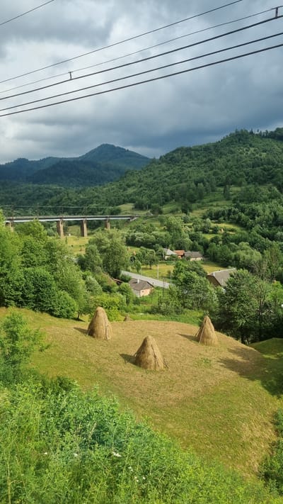 The image depicts a rural landscape with rolling green hills covered in dense forest. In the foreground, a mowed grassy slope features three tall, conical haystacks made of dried grass. A road winds through a small cluster of houses with visible roofs, nestled amongst trees and fields. Further in the distance, a multi-span railway bridge crosses a valley, with train tracks visible on top. The sky is overcast with dark, heavy clouds, suggesting potential rain. The overall scene conveys a tranquil, rustic atmosphere characteristic of a Ukrainian village. The image is taken from an elevated viewpoint, possibly from a train, as indicated by the overhead power lines in the upper portion of the frame.