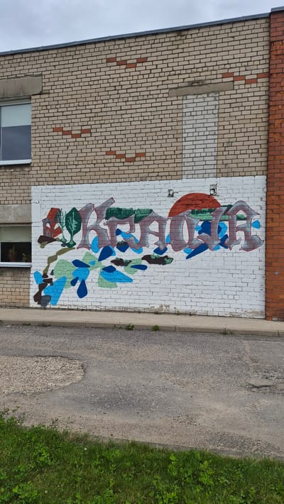 The image shows a vertical mural painted on a white brick wall of a building. The mural features stylized graffiti lettering in shades of brown, grey, and blue, with green and red accents. There are also abstract shapes and leaf-like forms interspersed within and around the lettering. To the left of the mural, the building's wall is constructed of light-colored bricks with some red brick accents and patches of concrete or plaster. A small section of a window is visible at the bottom left. To the right of the mural, there is a concrete curb and an asphalt surface, likely a road or sidewalk, bordered by a patch of green grass. The sky is overcast, suggesting it might be a cloudy day. The image appears to be taken from an elevated angle, looking down. There is no visible text in the image besides the graffiti lettering itself. The overall scene depicts an urban or semi-urban setting with public art on a building exterior.