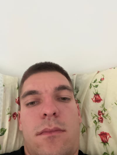 A close-up, low-angle, selfie-style photograph shows a person's face from the eyes down, with the head tilted upwards against a white ceiling. The person has short, dark hair, fair skin, and is wearing a black t-shirt. Their left eye is visible, and the right eye is partially obscured. Their nose and lips are also visible. The background consists of a white ceiling and a pillow with a floral pattern featuring red roses and green leaves. The image appears to have been taken indoors, possibly in a bedroom, with natural light. There is no identifiable information about the specific location in Bašť, Czechia.