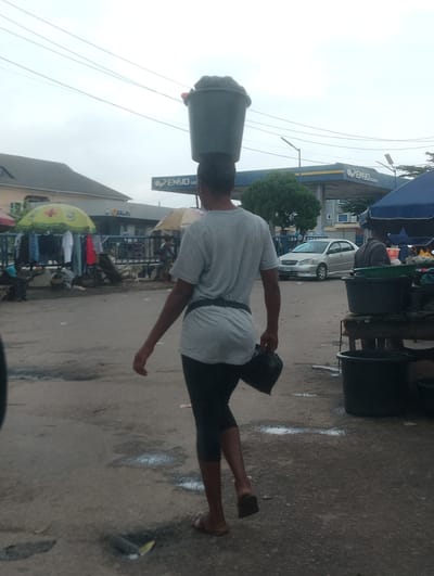 A person walks across a somewhat wet, dusty ground in Iju, Nigeria. They are carrying a large, dark-colored bucket balanced on their head. The person is wearing a light grey t-shirt, black leggings or tight pants, and sandals. A black bag is held in their right hand. The background features a gas station with the sign "ENYO" clearly visible. There are also market stalls with clothes displayed under umbrellas, a silver car, and several large black plastic containers. The sky is overcast, suggesting it might be a cloudy day or nearing evening. The overall scene appears to be a busy, everyday street market or roadside commerce area.