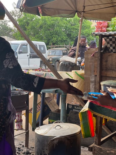 A market scene features a man on a motorcycle, smiling under a checkerboard awning. A white van and an older, green vehicle are visible behind him. In the foreground, a person's arm is raised in a greeting or gesture. Stacks of boxes, cooking pots, and various goods fill the frame, suggesting a bustling commercial environment. Colors, patterns, and objects are layered, creating a dense and vibrant composition.