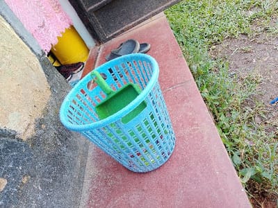 The scene, located in Sheywe ward, Kenya, depicts an outdoor area with a light blue, perforated plastic basket positioned on a reddish-brown ground surface. Inside the basket is a green plastic dustpan. To the left, a portion of a grey and tan textured wall is visible, partially obscuring a pink lace fabric draped over an unidentified object, with a yellow container visible beneath. A pair of dark-colored slip-on sandals rests near the wall, behind the basket. To the right of the basket, the reddish-brown surface transitions into an unkempt patch of green grass, weeds, and exposed soil. A small blue object is partially visible on the ground within the grassy area. No individuals are present; all objects are stationary.
