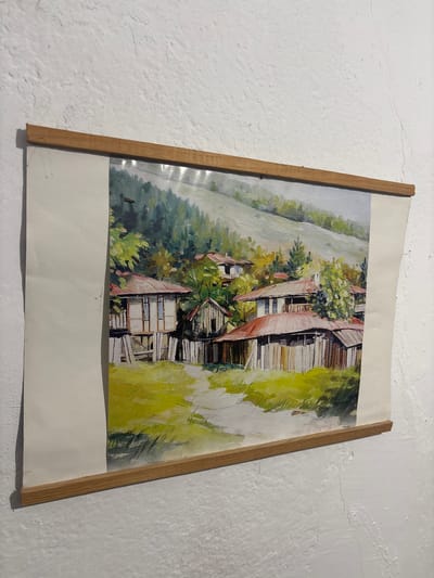 The image shows a painting of a village scene, likely in Shipkovo, Bulgaria, rendered in an impressionistic style with visible brushstrokes. The painting depicts traditional houses with red tile roofs and wooden structures, set against a backdrop of lush green trees and a gently sloping hillside. The foreground is a vibrant green field, suggesting a rural, possibly mountainous, environment. The painting is framed by two wooden slats at the top and bottom, and a light-colored border extends beyond the painted area. The artwork is displayed on a textured white wall.