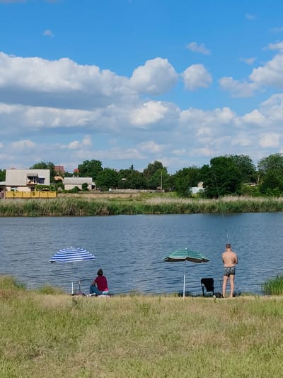 A wide-angle shot captures a calm, blue body of water bordering a grassy embankment. Two individuals are seen fishing by the water's edge. To the left, a person sits beneath a blue and white striped umbrella, facing the water. To the right, a shirtless individual wearing patterned swim trunks stands with a fishing rod, also facing the water. An umbrella with green and white stripes is set up near them, with a black chair beside it. The background reveals a line of trees and houses, suggesting a residential area. The sky above is a clear blue dotted with fluffy white clouds. The scene appears to be a recreational area within the city of Shyroka Dacha, Ukraine, on a sunny day.
