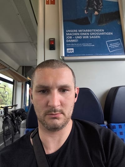 A man with a buzz cut and a beard, wearing a black t-shirt and black earbuds, is taking a selfie on a train. He is seated in the foreground, with two blue train seats visible behind him. To his left, two bicycles are visible through a window. A poster is displayed on the wall to his right, featuring text in German and an image of a person. The poster's text states "UNSERE MITARBEITENDEN MACHEN EINEN GROSSARTIGEN JOB – UND WIR SAGEN: DANKE!" which translates to "OUR EMPLOYEES DO A GREAT JOB – AND WE SAY: THANK YOU!". The poster also includes information about "UBB Usedomer Bäderbahn GmbH" and its location at "Am Bahnhof 1, 17424 Seebad Heringsdorf". This company operates in the region of Züssow, Germany.
