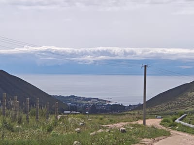 The image shows a landscape view with a body of water in the background, likely Issyk-Kul lake, near the city of Cholpon Ata, Kyrgyzstan. In the foreground, there is a dirt road curving to the right, with a grassy embankment on the left. A wooden fence with concrete posts runs along the embankment, separating it from the road. A single utility pole with wires is positioned near the right side of the frame. In the mid-ground, a town or city is visible along the shoreline of the water, with buildings and trees. The hills on either side of the frame frame the view of the water and the settlement. The sky is overcast with scattered clouds. There are no people visible in the image.