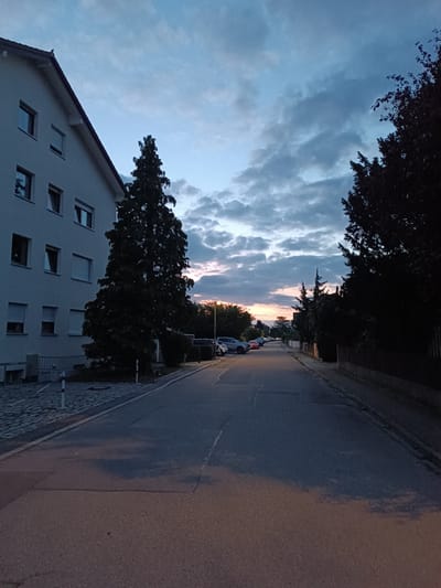 The image depicts a street scene in Germering, Germany, at dusk. A multi-story building with several windows and light-colored siding is visible on the left side of the frame. In front of the building, a paved walkway leads to the street, with a metal fence and some small bollards. A tall, dark evergreen tree stands prominently between the building and the street.
The street itself is asphalt, with a faded white line marking its center. Several cars are parked along the right side of the street, and a few more are visible further down the road on the left. The street is lined with trees and vegetation. A wooden fence runs along the right edge of the road.
The sky is partly cloudy, with a gradient of colors indicating the setting sun. The clouds are dark and textured, contrasting with the lighter blue of the sky. The overall lighting suggests a time shortly after sunset. The scene is quiet and appears to be residential.