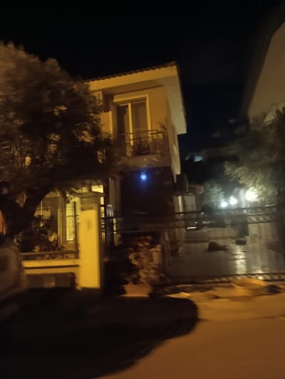 This is a nighttime, slightly blurry image of a multi-story building in Urla, Turkey. The building has a light-colored facade with a red-tiled roof. There is a balcony with a railing on the second floor. A large tree with dark foliage partially obscures the left side of the building. The ground in front of the building appears to be paved and wet, reflecting the light. There are several outdoor lights visible, some of which are blurred. A dark, shadowy area in the foreground could be a lawn or a dark pathway. The sky is dark, indicating nighttime. The overall impression is of a residential building at night. There are no visible people or text in the image.