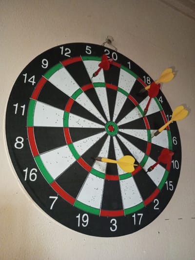 A dartboard hangs on a light-colored wall. The board is black and white with green and red rings, numbered 1 through 20. Five darts are stuck in the board. A red dart is in the "20" section. Two yellow darts are in the "6" and "7" sections. Two more red darts are in the "4" and "10" sections.
