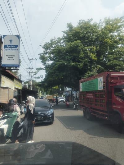 The image shows a street scene in Keudematangglumpangdua, Indonesia. A red truck carrying LPG gas cylinders is parked on the right side of the road. The truck has signage indicating it is for PT Sadaijuta Putra Perkasa, a supplier of subsidized LPG for low-income families. A pharmacy sign for "Apotek Qiyya Farma" is visible on the left, with text indicating it accepts doctor's prescriptions. Several motorcycles are on the road, with two individuals on one motorcycle in the foreground wearing headscarves. A black Toyota car with license plate "BL 1097 ZR" is stopped in traffic. The environment appears to be an urban area with trees, overhead electrical wires, and various buildings lining the street. The sky is bright and overcast, suggesting daytime.
