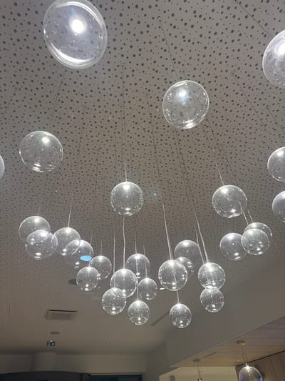 The image displays an interior space located in Marcq-en-Barœul, France, featuring an overhead lighting installation. Approximately forty clear, spherical glass pendant lights are suspended at varying heights from a light-colored ceiling perforated with a dense pattern of small circular holes. Each glass sphere encases an illuminated light source, visible as a bright internal point, and the spheres exhibit reflective surfaces. Thin suspension wires connect the individual light fixtures to the ceiling. In the lower left portion of the frame, a dark rectangular opening is partially visible beneath the ceiling, adjacent to a light-colored rectangular air vent. A small green square sign with a white pictogram is affixed to the ceiling. The lower right segment of the image reveals a partial view of a light-colored structural beam or wall, beneath which additional, less distinct lighting elements or reflections are present. No individuals or active interactions are depicted.