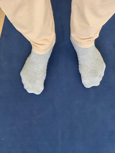 This is a top-down medium shot of two feet wearing light gray socks, standing on a dark blue mat. The mat has a subtle textured pattern. The feet are positioned slightly apart and angled away from each other. Light peach-colored pants are visible covering the lower legs. The lighting suggests an indoor setting, likely during the daytime. There is no visible text in the image. The context provided places the scene in Gossau, Switzerland.