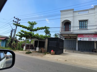 The image shows a street scene in Panteepanah, Indonesia. On the right side of the frame, there is a two-story white building with a storefront. The storefront has two businesses, "Brother Textile" and "Fahmi Optical," with closed grey roller shutter doors. "Fahmi Optical" has a sign advertising eyeglasses and prices.

To the left of the building is a small, black rectangular structure with a serving window, potentially a coffee bar or food stall labeled "Si Coffee Bar". Further left are some overgrown plants, a makeshift shelter, and a parked motorbike. A wooden utility pole with visible electrical lines is on the left side of the frame.

The foreground features a two-lane asphalt road, with the left side of a white vehicle visible in the side mirror of another vehicle, suggesting the photo was taken from a moving car. The sky is blue with scattered clouds.