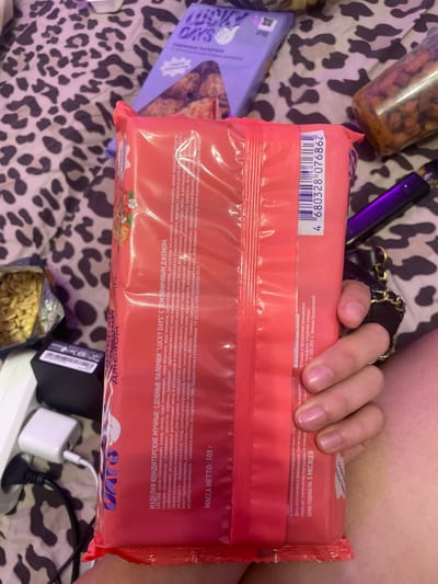 The image displays a close-up view of a hand holding a red package of "Lucky Days" strawberry-flavored confectionery sticks. The background consists of a patterned blanket and other items, including a charging device and a vape pen. The red package has text in Russian, including the product name, ingredients, and weight (308g). There is no visible information indicating the location in Zmeinyi, Russia.