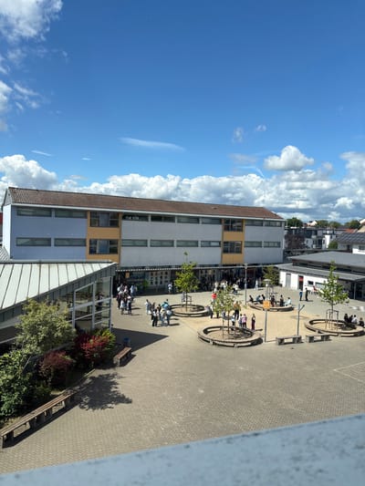 The image depicts an outdoor courtyard area of a school in Groß-Gerau, Germany, under a bright blue sky with scattered white clouds. The foreground shows a light-colored railing, suggesting the photo was taken from an elevated position. The central area is paved with light grey interlocking bricks and features several circular wooden benches surrounding small trees. In the background, a multi-story school building with a brown tiled roof dominates the scene. The building has sections painted in light blue, orange, and white, with numerous windows. To the right of the main building, a smaller, single-story building with a grey roof is visible.

Scattered throughout the courtyard are approximately 30-40 individuals, appearing to be students and possibly some adults, in various states of interaction. Some are standing in small groups, others are walking, and a few are seated on the benches. The individuals are dressed in casual clothing, indicating a typical school day.

To the left of the main building, a modern glass-fronted structure with a sloped metal roof is partially visible, adjacent to a green area with trees and bushes. The overall atmosphere is that of a bustling school environment on a sunny day. The presence of the school building, the paved courtyard with recreational seating, and the gathering of people strongly suggest this is a school campus.