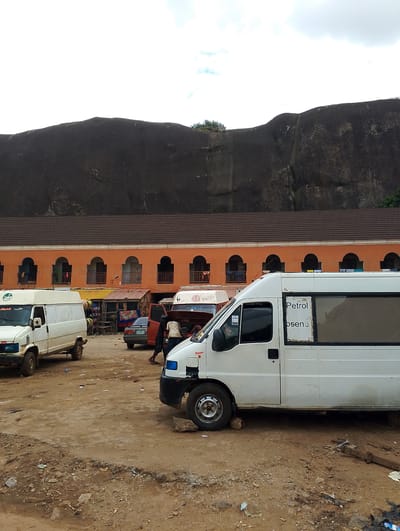The image depicts a street scene in Oba-Ile, Nigeria, with a prominent large rock formation in the background. In the mid-ground, an orange building with arched windows and a tiled roof runs horizontally across the frame. In front of the building, several vehicles are visible, including white vans and a car. One white van in the foreground has the word "Petrol" and some illegible text visible on its side. People are seen interacting with the vehicles, with one person appearing to work on the open hood of a vehicle. The ground is unpaved and appears dusty and littered with debris. The sky is overcast.