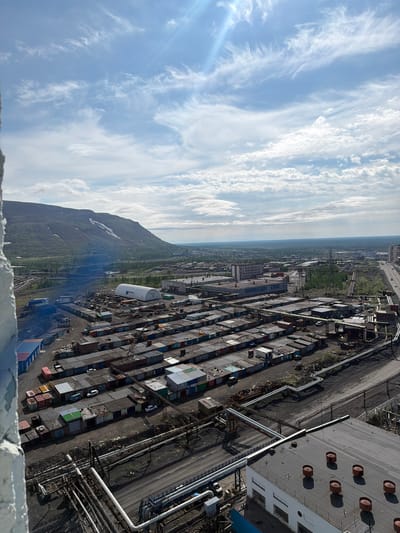 The image shows an aerial view of an industrial area in Norilsk, Russia. In the foreground, there is a large building with a flat roof and several circular vents. Surrounding the building are numerous pipes and industrial infrastructure.
Further into the image, there is a dense area of small, uniformly constructed structures, likely garages or storage units, arranged in neat rows. Beyond these, more industrial buildings are visible, including a large white arched structure. In the background, a vast, relatively flat landscape stretches towards the horizon, with some vegetation and power lines. A prominent, green, mountainous terrain with patches of snow is visible to the left, dominating the background. The sky is partly cloudy with a bright sun. The scene appears to be a snapshot of a working industrial city, possibly on a summer day given the clear sky and presence of snow on the distant mountains. There are no visible people in the image.