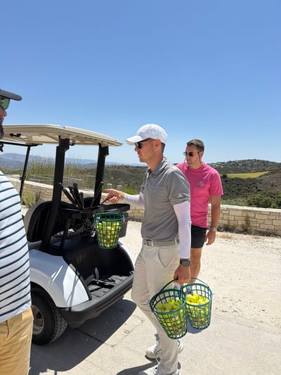 Three men are in Tsada, Cyprus, standing near a white golf cart on a sunny day. The man on the left, wearing a green and white striped shirt and tan pants, is partially visible from the side. In the center, a man in a grey polo shirt, white cap, and light-colored pants, is holding two green baskets filled with yellow golf balls. He gestures towards the golf cart with his right hand. Behind him, a man in a bright pink t-shirt and dark shorts, wearing sunglasses, stands facing away from the camera. The golf cart has several clubs in a holder. The background shows a dry, hilly landscape with some buildings visible on the slopes under a clear blue sky.