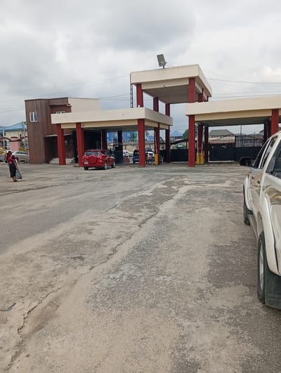 The image depicts an outdoor scene at what appears to be a gated entrance or facility in Eliozu, Nigeria. In the foreground, the right side of a white pickup truck is visible. The ground is paved with asphalt and shows signs of wear and tear, with cracks and uneven patches.

In the mid-ground, a red SUV is parked. To the left, a woman in a black dress and headscarf is walking away from the camera, carrying a black plastic bag. Behind her, a light-colored sedan is parked.

Dominating the mid-ground and background are structures with red pillars supporting cream-colored roofs, resembling an open-sided canopy or entryway. Beyond these structures, a black metal gate is visible, with buildings and more structures behind it. The sky is overcast with gray clouds. The overall impression is of a functional, utilitarian space, likely a commercial or administrative area within the city.