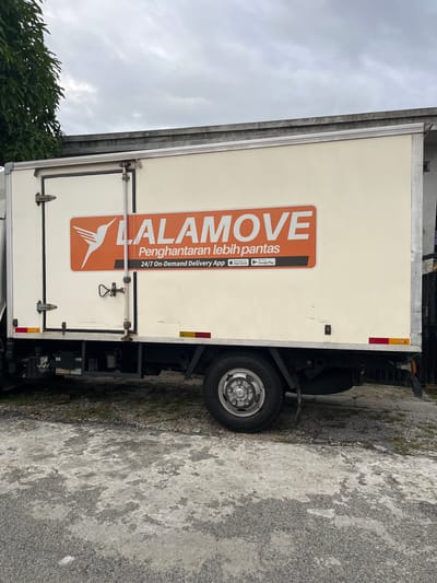 The image shows the side of a Lalamove delivery truck parked on an asphalt surface. The truck is white with an orange and white Lalamove logo and text on the side. The text includes "Penghantaran lebih pantas" and mentions the 24/7 on-demand delivery app. The truck is parked next to a curb and some vegetation is visible in the lower left corner. The sky is overcast. The location is the city of Ipoh, Malaysia.