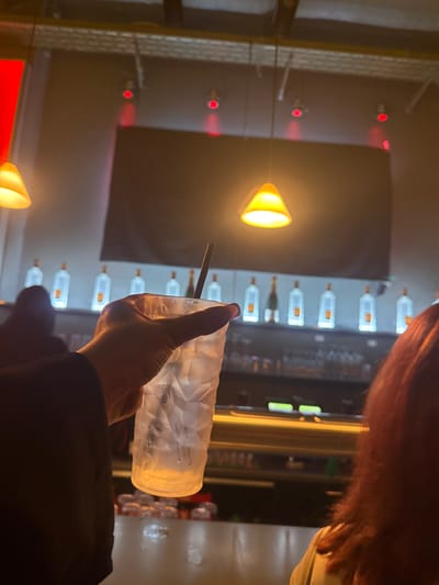 Inside a dimly lit bar, a person holds a textured glass filled with ice and a straw. Behind them, a bar is lined with illuminated liquor bottles. Three cone-shaped lights hang, casting warm glows, while red lights dot a mirrored surface on the left. The person wears a dark top and has reddish hair. The photograph has been rotated, giving a slightly disorienting perspective.