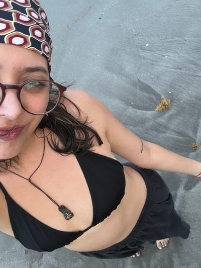 A woman is lying on a gray surface, possibly sand, in Mata de Uva, Mexico. She wears a black bikini top and dark, loose-fitting pants. The woman also has a patterned headscarf and glasses. Small clusters of dry plant matter are scattered on the ground.