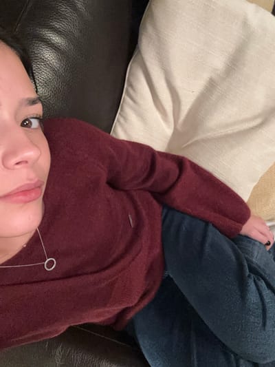 A young adult individual, appearing female, is depicted in a partial, slightly high-angle indoor shot, reclining on dark brown or black leather furniture. The subject's left facial profile, including a dark eye, nose, and lips with light pink lipstick, is visible, with her gaze directed towards the camera. She wears a long-sleeved, maroon or burgundy knit sweater, which features a small, vertical silver-colored tag on the upper left chest area, and dark blue denim jeans. A delicate silver-toned chain necklace with a small, open circular pendant is worn around her neck. Her left hand, partially visible, shows black nail polish. To her right, an off-white textured throw pillow is positioned, with a segment of beige fabric beyond it. This scene takes place indoors in the city of Huasca de Ocampo, Mexico.