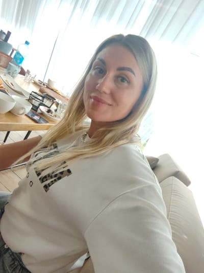 The image shows a woman taking a selfie in a brightly lit room, possibly a cafe or restaurant, with white curtains in the background and a table with food and drinks in front of her. The woman has blonde hair and is wearing a white t-shirt with a leopard print design. The table has plates with remnants of food, cups, a bottle of water, and a metal wire basket. The presence of a sofa is visible in the lower right corner of the image. The image does not provide any specific visual cues to determine the location within the city of Amursk, Russia.