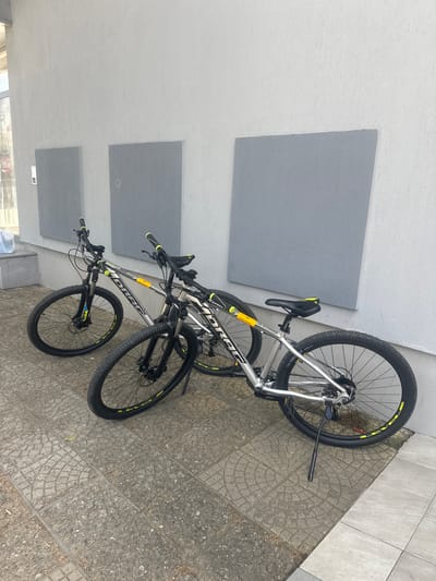 Two silver and black mountain bikes are parked on a sidewalk next to a textured light grey wall in Ovoshtnik, Bulgaria. The bikes are positioned parallel to each other, with the front wheels slightly turned outwards. Both bikes have black tires with yellow accents, black handlebars with yellow grips, and black seats with yellow accents. One bike has a yellow sticker on its frame. The sidewalk is paved with interlocking square tiles, some of which are decorated with a circular pattern. The wall behind the bikes has two large, square grey panels mounted on it. To the left of the bikes, a narrow concrete step leads upwards. A blue plastic bag is visible on the step. The scene appears to be outdoors, possibly in front of a building.