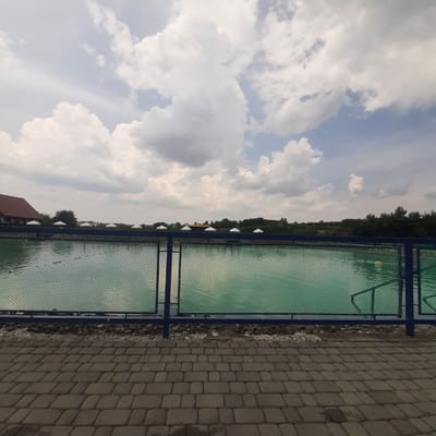 The image depicts a man-made pond in Dubrivka, Ukraine, likely an aquaculture or fishing facility, based on the presence of what appear to be fish cages or netting in the water. A dark blue metal fence with green mesh separates the viewer from the pond. The pond's water is a vibrant turquoise color, reflecting the cloudy sky above. Along the far bank of the pond, several white canopy tents are visible, suggesting an area for visitors or vendors. Behind the tents, a dense line of trees forms the horizon. To the left, a rustic building with a red roof is partially visible. The foreground consists of a paved walkway made of interlocking gray bricks. The overall scene is naturalistic, with the pond, vegetation, and sky dominating the landscape. There are no people visible in the image.