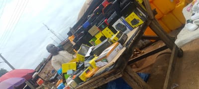 The image shows a man standing behind a stall laden with various electronic devices and accessories, likely in a marketplace in Baissa, Nigeria. The stall features a wide assortment of items, including portable speakers, headphones, chargers, and other electronic gadgets, many of which are in their original packaging. The packaging is predominantly yellow and black, with some items also in white.

The man, dressed in a light-colored traditional outfit, appears to be a vendor attending to his stall. He is looking slightly upwards and to the right, his expression neutral. The stall itself is constructed from rough wooden planks, suggesting a rustic market setting. Behind the stall, large yellow plastic containers are stacked, and further back, a building with a corrugated iron roof is visible. Power lines and two tall communication towers are prominent in the background, indicating an urban or semi-urban environment.

The sky is overcast, suggesting a cloudy day, possibly late morning or afternoon given the lighting. The ground is a mix of dirt and sand. To the left of the stall, a red and a purple umbrella are partially visible, adding to the market atmosphere. A few pairs of white shoes are also visible on a surface to the right.

Visible text on the packaging includes "Super Bass", "PORTABLE SPEAKER", "LED 100W WHITE", and "LN-BSGT BLUETOOTH". The overall impression is a busy, open-air marketplace where electronics are being sold.