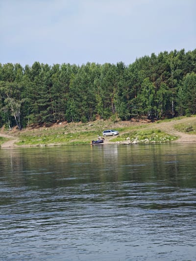 A wide, calm river flows in the foreground, reflecting the trees and sky. The riverbank on the far side is lined with a dense forest of pine and birch trees. Two silver-colored cars are parked on the grassy bank near the edge of the forest. A small inflatable boat with two people is visible on the water, close to the shore. The sky is clear and pale blue. The scene depicts a tranquil natural setting on a sunny day.