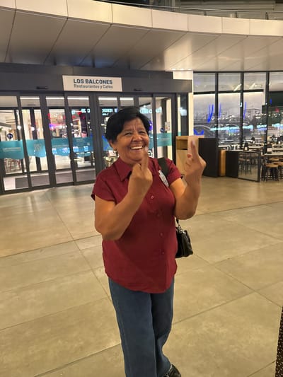 An older woman with dark hair and a red shirt is standing in front of the entrance of "LOS BALCONES Restaurantes y Cafés" in Alcorcón, Spain. She is smiling broadly and holding up both middle fingers in a defiant gesture. The entrance is marked by large glass doors and a sign above them. The background shows a modern interior with tables and chairs visible through the glass. The floor is tiled and the lighting appears to be artificial, suggesting it is indoors or covered.