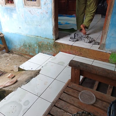 The scene is located in Pameungpeuk, Indonesia, specifically at the coordinates -7.641 latitude and 107.728 longitude. The image depicts a partially covered exterior staircase with white tile steps leading up to a wooden doorframe.  The staircase is bordered on the left by a blue wall with green and brown moss growing on the lower portion. A small wooden bench rests on the top step of the stairs,  and a gray fabric item is lying on the floor inside the doorway.  A pair of brown sandals and a brown wooden bench rest on the lower steps. The person whose legs are visible in the image is wearing a pair of green pants and has exited the doorway to the left. The scene appears to be partially covered in dirt and debris.