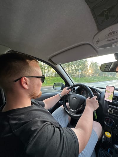 A medium shot shows a Caucasian man with light skin, reddish-brown hair, and a goatee, wearing sunglasses and a black t-shirt, driving a car. The man's left hand is on the steering wheel, and his right hand is also on the wheel. He is looking forward, presumably at the road ahead. The interior of the car is visible, including the dashboard, steering wheel, and a phone mounted on the windshield displaying a navigation app. The exterior of the car shows a road with trees and sunlight in the background, suggesting an outdoor, possibly rural setting. The image is taken from inside the car, looking towards the driver. The scene suggests a person driving on a road in Opalikha, Russia, with the sun setting or rising, casting a warm glow.