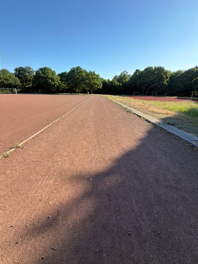 The image depicts a track and field area under a clear blue sky, likely in the city of Dorsten, Germany. The foreground features a reddish-brown cinder track, divided by a thin concrete line with some sparse grass growing along its edge. The track appears to be curving slightly away from the viewer. In the middle ground, a line of mature green trees forms a dense border. Beyond the trees, a red sports field, possibly a soccer field, is visible. A lone person, seen from behind and at a distance, is walking on the track near the tree line. The lighting suggests it is either early morning or late afternoon, casting long shadows across the track. The overall impression is one of an outdoor sports facility bathed in natural light.