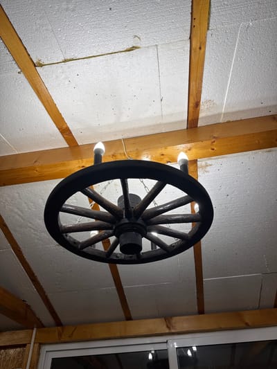 The image shows a wagon wheel chandelier hanging from a wooden beam ceiling in what appears to be an indoor setting. The ceiling is constructed with white, textured panels and reinforced with wooden beams. The chandelier itself is made of dark wood and has several light bulbs protruding from its spokes. The lighting is dim, casting shadows and highlighting the rustic design of the chandelier and ceiling. The background shows a sliver of a white window frame and some reflective surfaces, suggesting this could be a home or a commercial establishment. There are no people or animals visible in the image, and no specific details about the location in Mineralni Bani, Bulgaria are provided or inferable from the visual content.