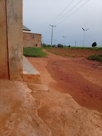 The image captures a scene on a dirt road in Ban, Nigeria. In the foreground, a textured concrete structure, possibly part of a building's foundation or a raised platform, takes up the left side of the frame. The ground is a reddish-brown dirt, with areas that appear wet or muddy.

Further into the scene, a dirt road curves into the distance. On the right side of the road, there's a field with sparse green vegetation. On the left side of the road, a building constructed from brick is visible, with a green-roofed structure behind it. A patch of green grass is in front of the brick building.

The background is characterized by an overcast sky and a flat, open landscape. Several tall utility poles with wires stretch across the sky. Three modern streetlights are visible along the road, suggesting the area has some infrastructure. A lone tree stands on the right side of the frame, and another clump of trees is further in the distance. A vehicle is parked or driving on the road in the far distance. The overall impression is of a rural or semi-urban area undergoing development, with a mix of existing structures and new infrastructure.